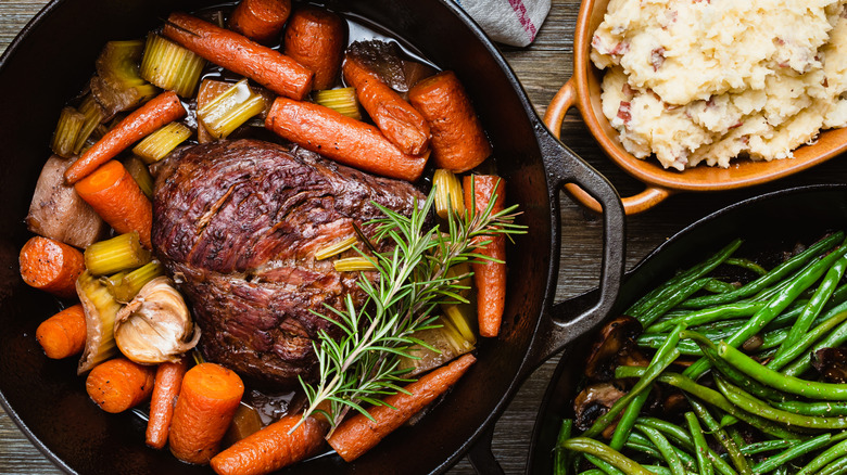 Cast iron with pot roast, veggies, and rosemary next to sides of mashed potatoes and green beans