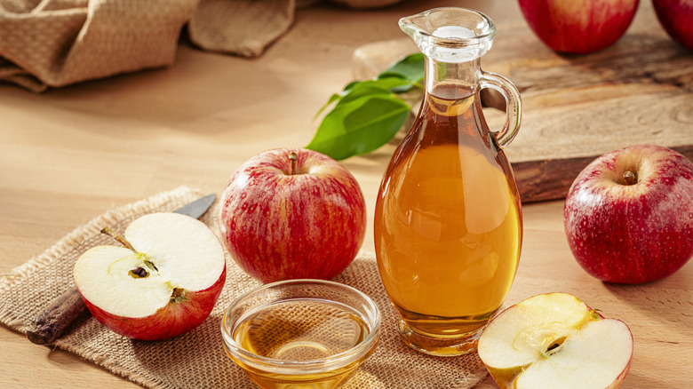 A bottle of brown liquid surrounded by apples and a small bowl of brown liquid
