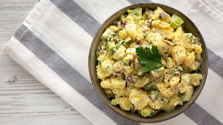 Top-down view of potato salad with chopped celery, red onion, and parsley in a bowl over a neutral-striped dish cloth