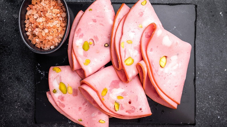 Folded slices of mortadella next to a small bowl of Himalayan pink salt on a black serving board.