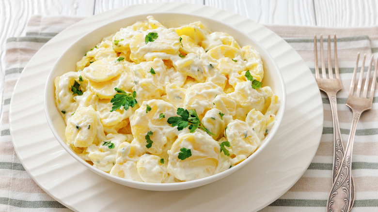 A white bowl of creamy potato salad on a charger plate next to forks