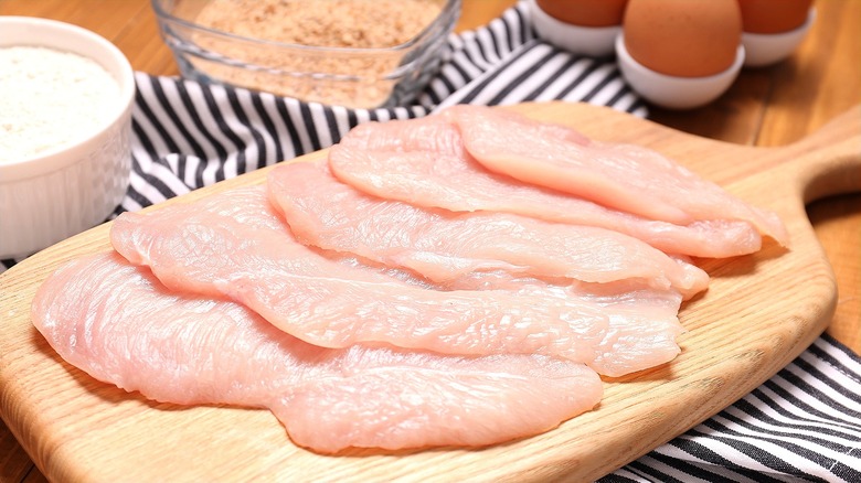Raw chicken cutlets on a wooden serving board surrounded by whole eggs, flour, and breadcrumbs