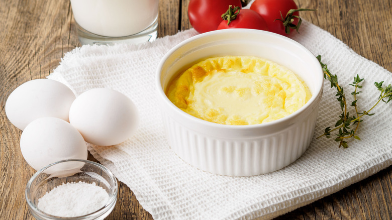 A white ramekin filled with an egg scramble, surrounded by whole eggs, tomatoes, salt, and a sprig of thyme.
