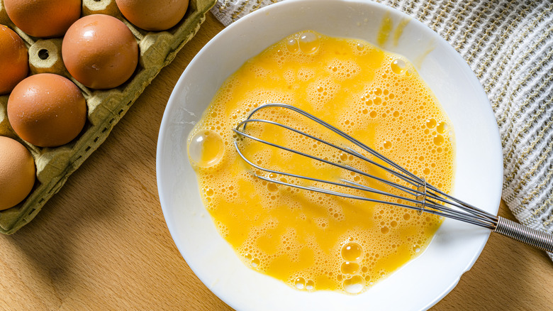 A bowl of freshly whisked eggs, a whisk on top, a carton of eggs next to it.