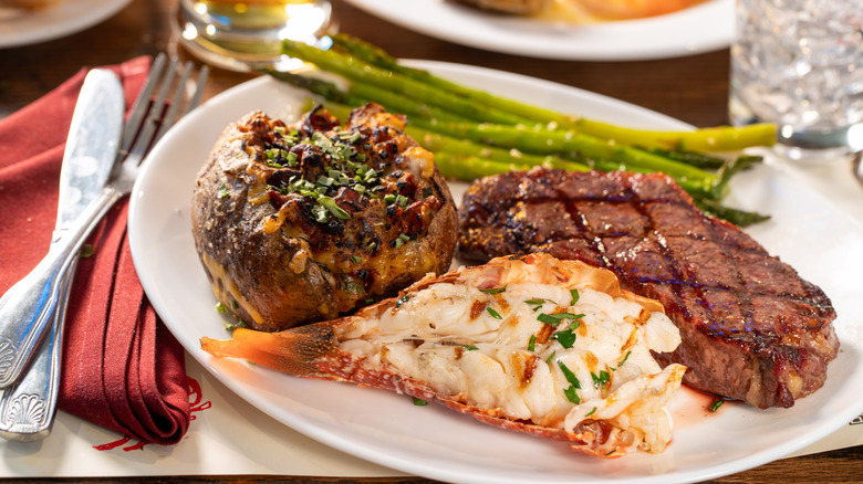 Steak, lobster tail, loaded baked potato, and asparagus on a white plate