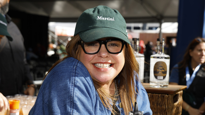 Rachael Ray smiling and wearing black glasses and a green hat that reads 