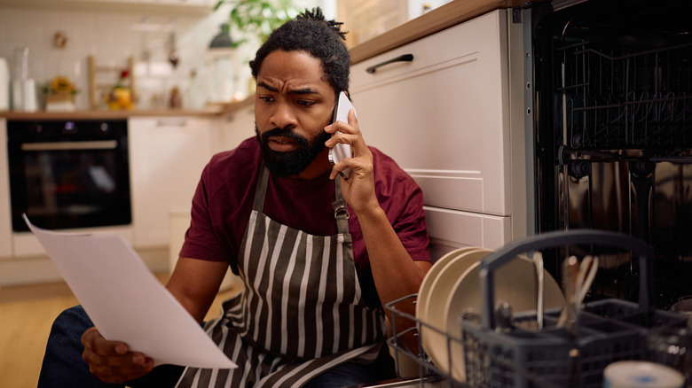 Man sitting beside a broken, open dishwasher calling for repairs