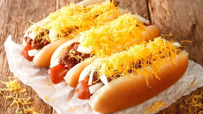 Three chili cheese dogs with chopped onions over a piece of white parchment paper on a wooden table