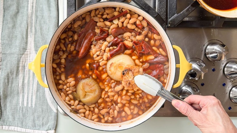 Removing a head of charred garlic from a pot of white beans cooking with onions and chiles