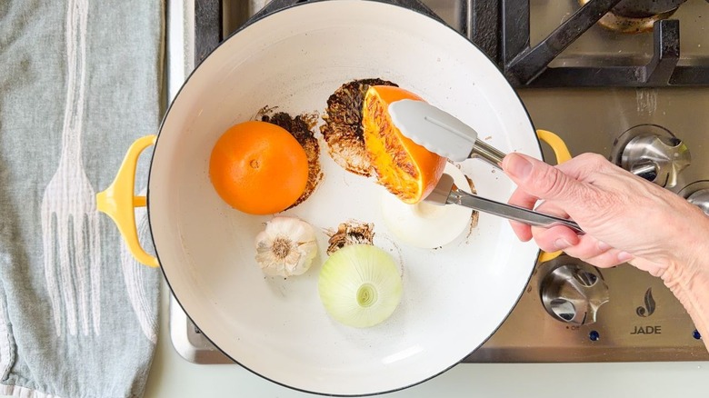 Lifting a caramelized orange half with tongs from a Dutch oven with white onion and garlic