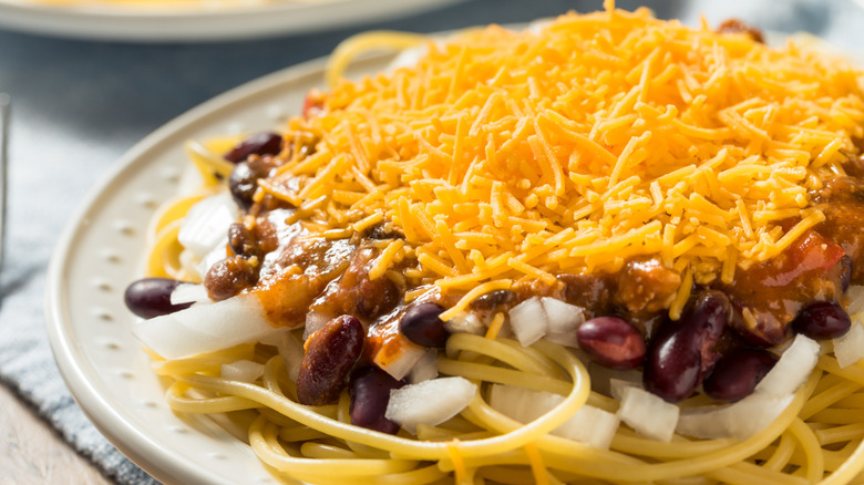A white plate topped with spaghetti, onions, chili, and shredded cheese.