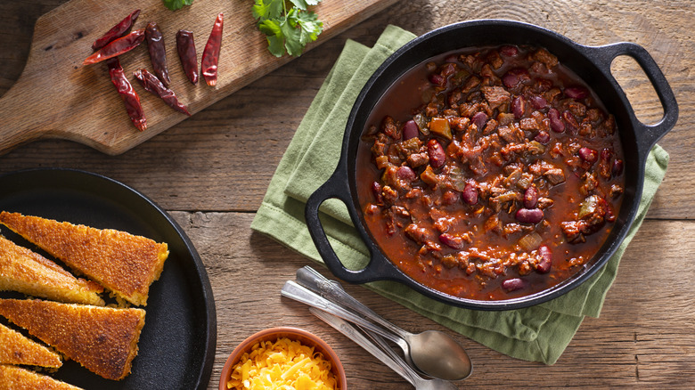 A pot of chili next to chilis, cilantro, cheese, and cornbread