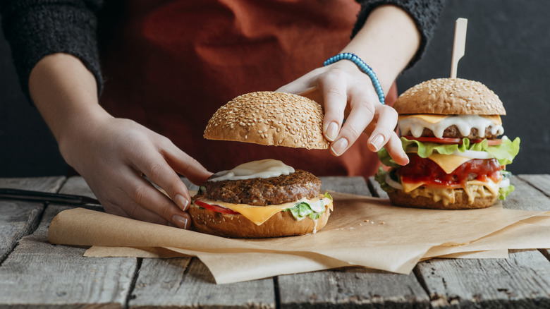 cropped view of girl in apron making homemade cheeseburgers on baking paper on wooden tabletop