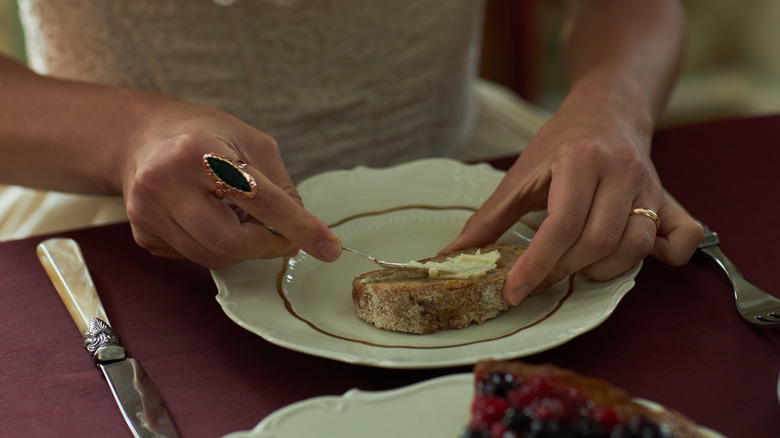 Woman spreading butter on toast in historical dining setup.
