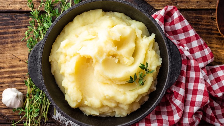 Mashed potatoes in a cast iron crock on a wooden table with herbs, garlic, and a red and white checkered napkin.