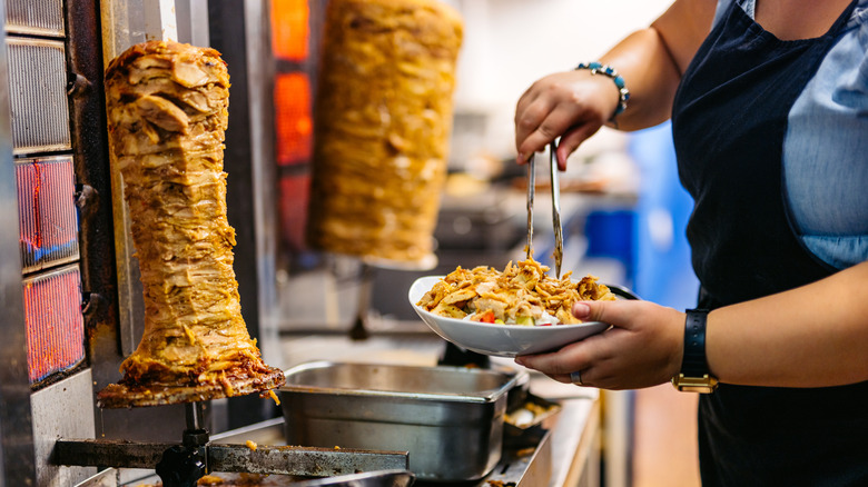 Woman worker in restaurant kitchen using tongs to put shawarma meat from spit onto a plate