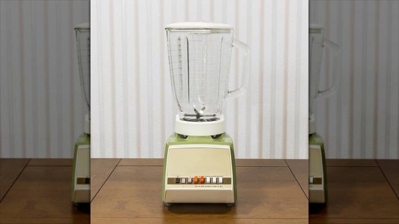 An avocado green vintage blender sitting on a table with striped wallpaper behind it