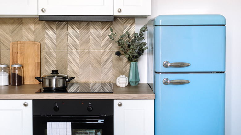Front view of stylish interior design in kitchen. White furniture, tiled wall, built-in appliances, blue retro refrigerator in apartment. Kitchenware and home decor on wooden countertop