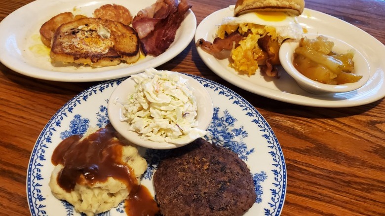 Plates of hamburger steak, eggs in the basket, and breakfast burger from Cracker Barrel arranged on a table.