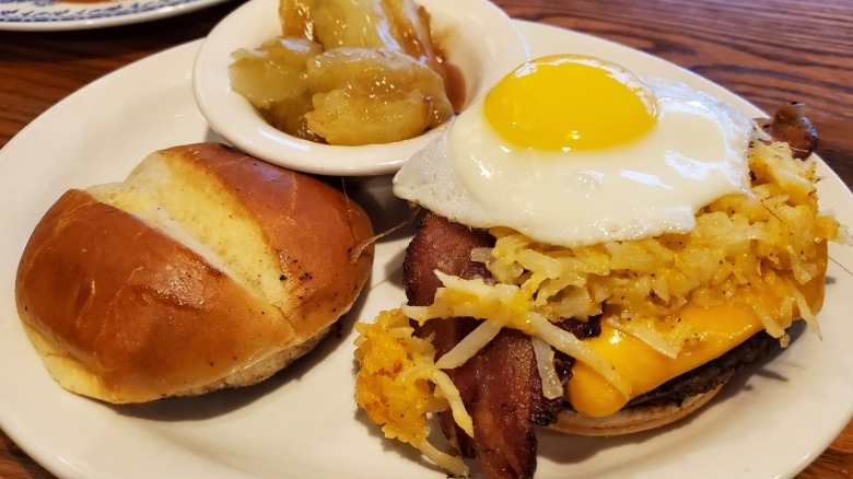 Cracker Barrel breakfast burger with the top bun lying on the side so you can see the sunny side up egg and has browns atop the burger plus a side of fried cinnamon applesde