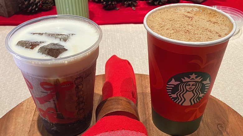 Two Starbucks holiday beverages sitting side by side on wooden surface with red rolled napkin between them