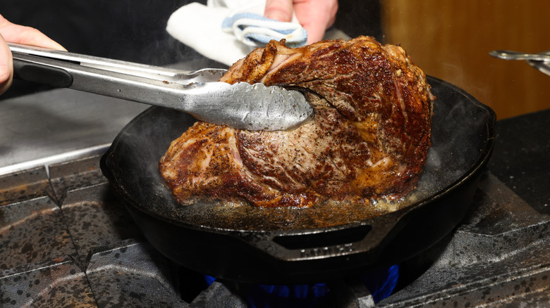 A cook flipping a ribeye with tongs in a cast iron skillet