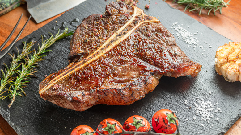 A grilled T-bone steak on a black slate served along with tomatoes, garlic, and fresh herbs