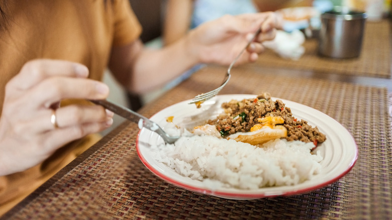 Person eating rice and meat off of a dish with a fork and spoon