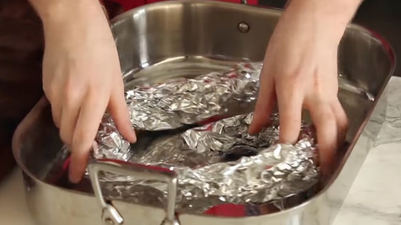 Hands placing a DIY roasting rack made of aluminum foil in the bottom of a roasting pan