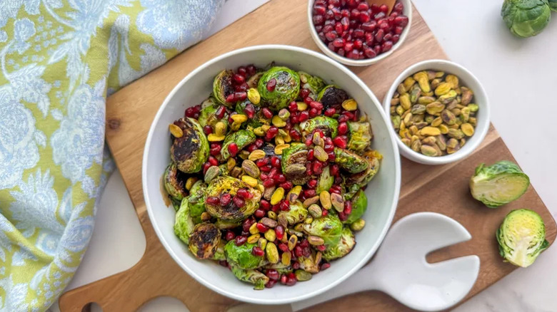 Crispy fried Brussels sprouts with pomegranate seeds next to bowls of pistachios, pomegranate seeds, and sliced Brussels sprouts.