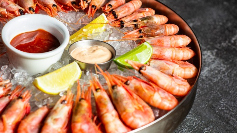 Royal red shrimp with the heads still on arranged in a circle on ice served with lemon, lime, and dipping sauce.
