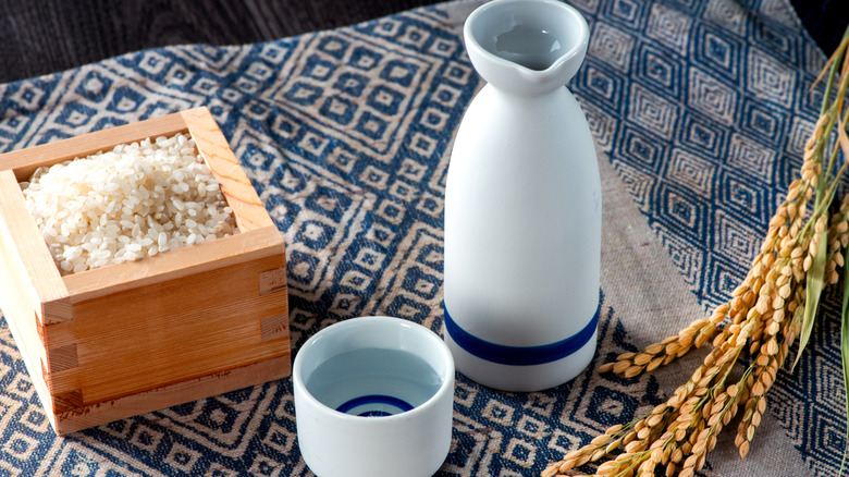 A porcelain sake bottle and matching cup, both white with a blue stripe at the bottom, next to a wood bowl filled with white rice and a sprig of harvested rice
