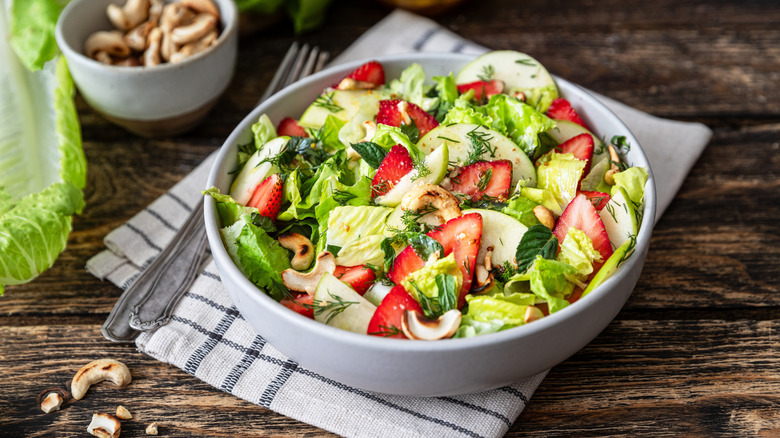 A fresh tossed salad with romaine lettuce, cucumbers, strawberries, and herbs, nuts in the background.