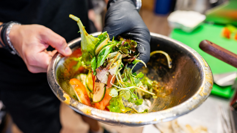 A chef with protective gloves hand-tossing a salad in a stainless steel bowl.