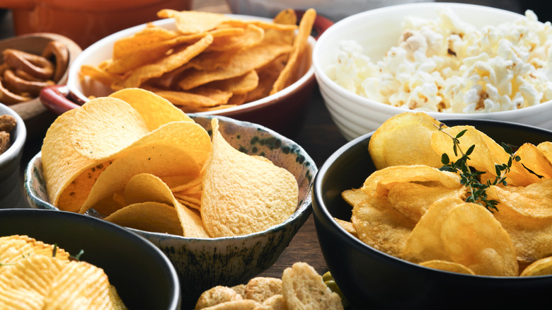 A variety of bowled filled with crunchy snacks like different kinds of chips, pretzels, and popcorn.