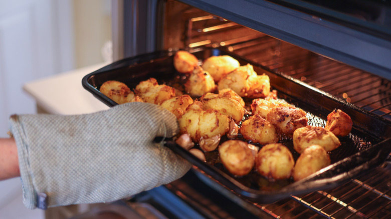 A person's hand in an oven mitt is seen removing roast potatoes from an oven.