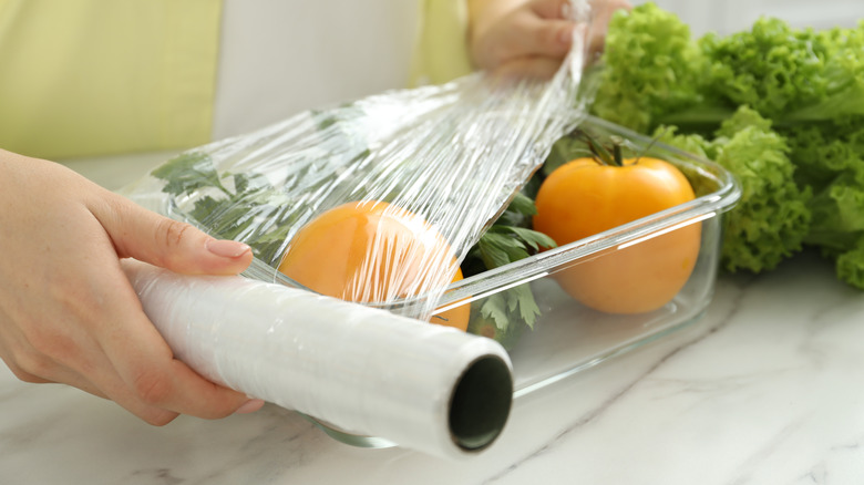Woman putting plastic food wrap over glass container with fresh vegetables at white marble table, closeup