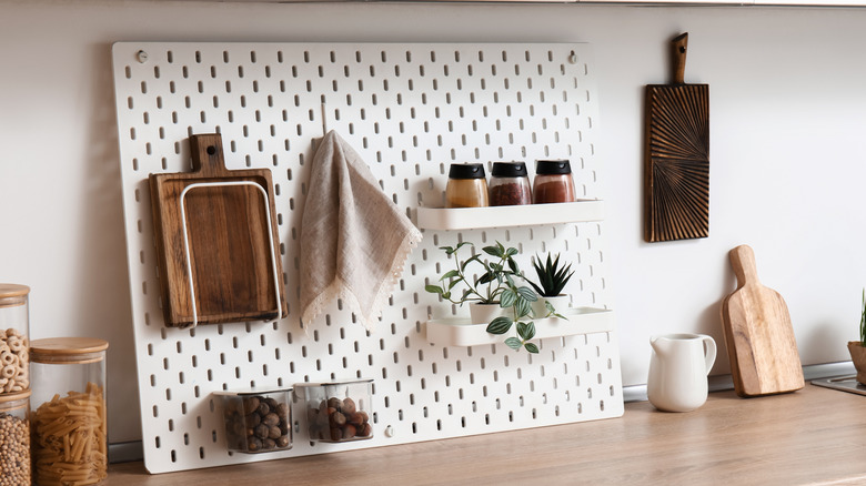 A white pegboard holding a wooden chopping board, a towel, several spices, and even potted plants