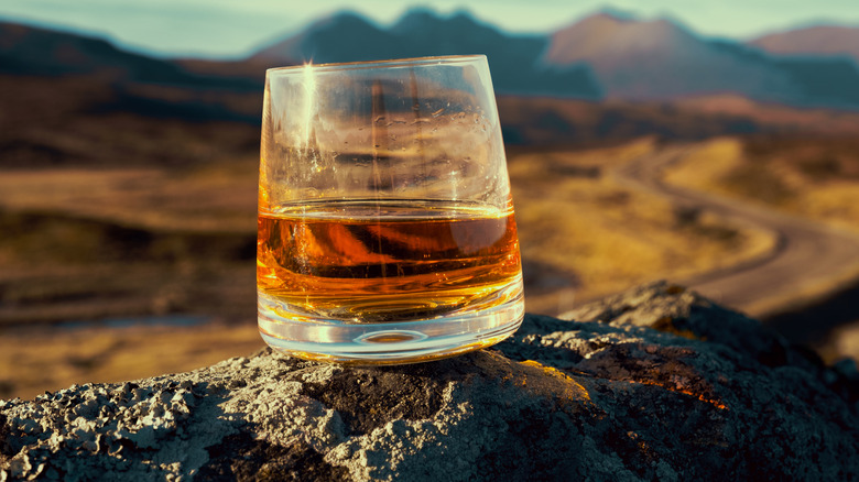 A glass of whiskey sits on rock with a landscape in the background of what could be Scotland or Ireland.