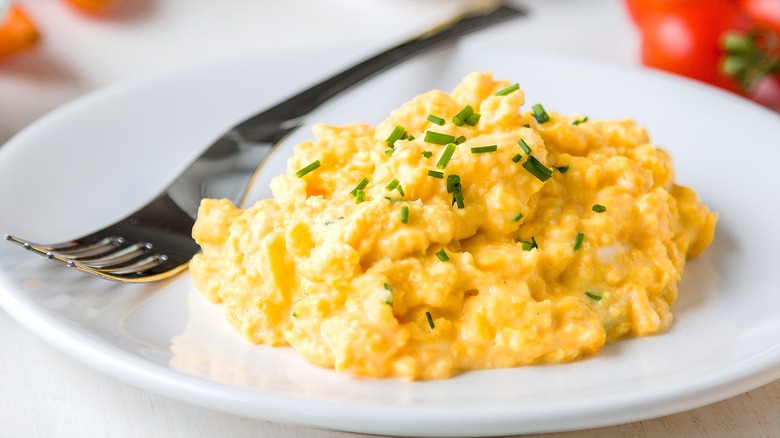 Soft scrambled eggs with chopped chives on a white plate with a metal fork