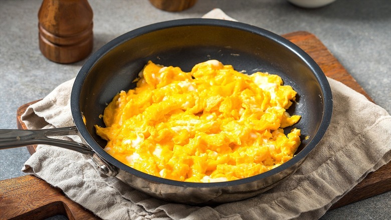 Scrambled eggs in a dark-coated frying pan on a folded cloth napkin over a wooden serving board