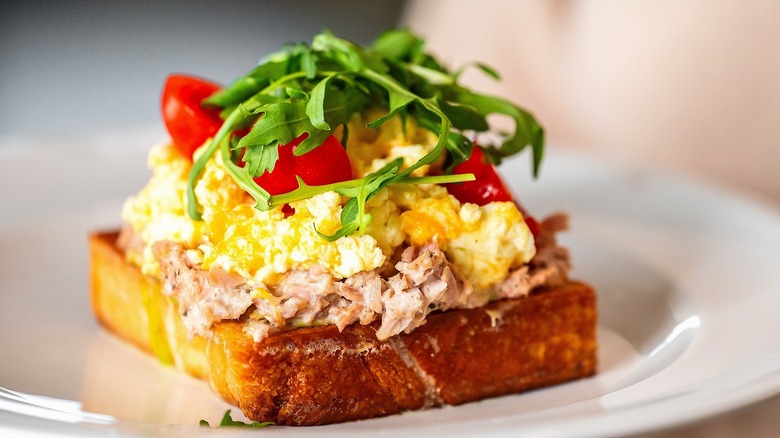 Toast topped with tuna salad, scrambled eggs, sliced cherry tomatoes, and arugula on a white plate.