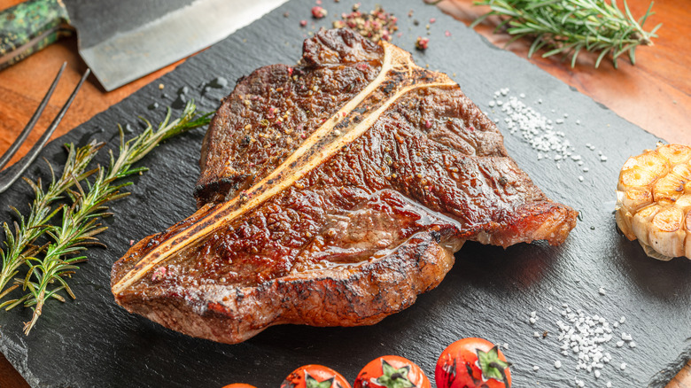 Cooked T-bone steak on a slate cutting board with herbs, garlic, tomatoes, and seasonings