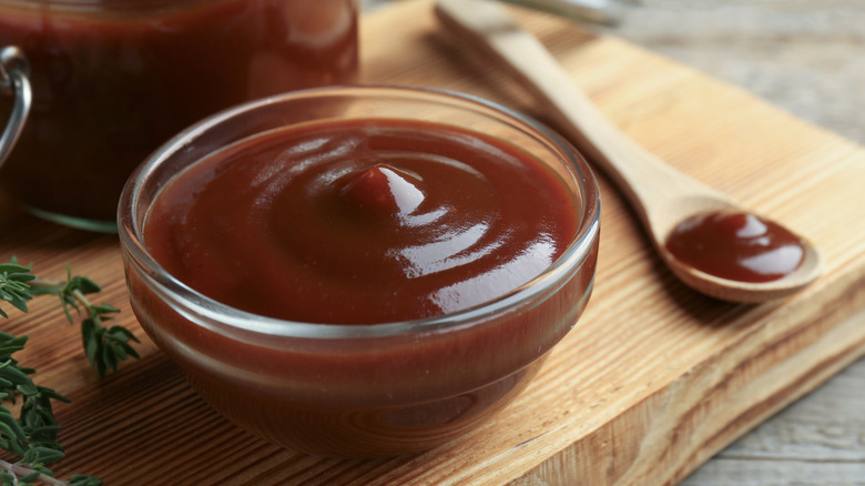Glass bowl with barbecue sauce sitting on a wooden plank next to a spoon full of barbecue sauce