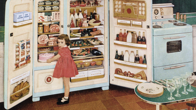 A little girl in 1955 opening a refrigerator extremely full of ingredients.