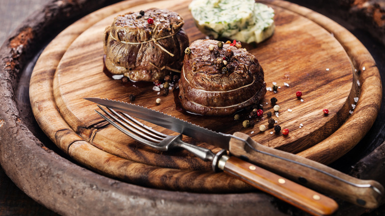 Point of view shot of a fork and serrated steak knife resting on a cutting board with a round steak