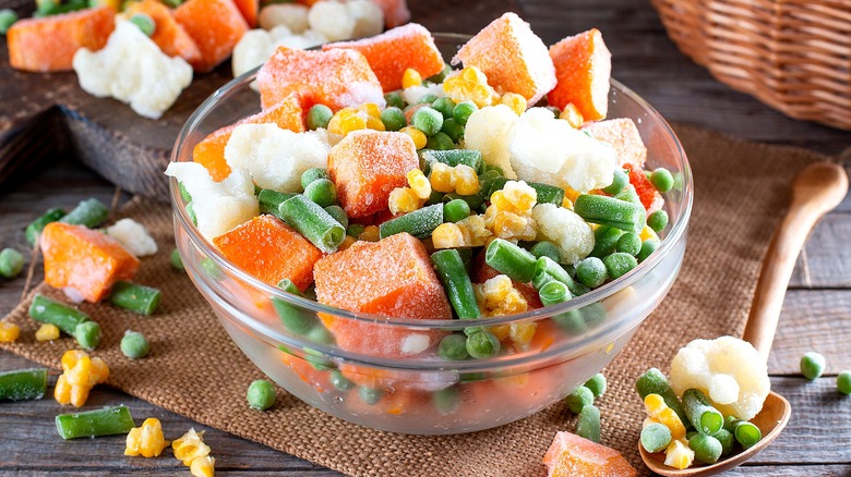 Frozen cut sweet potatoes, green beans, peas, and carrots in a clear bowl on a rattan cloth