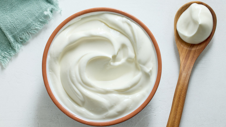 A bowl of sour cream is beside a wooden spoon full.