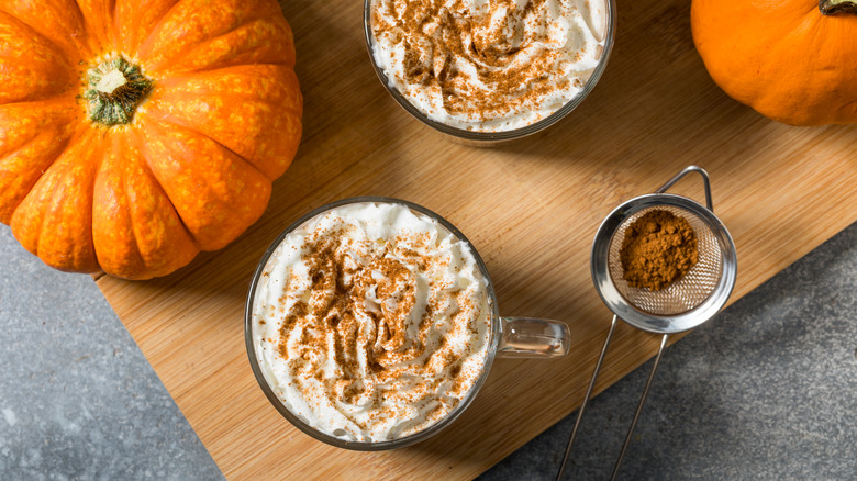 Top-down view of two pumpkin spice lattes on a wooden cutting board next to pumpkins and a small sifter of cinnamon
