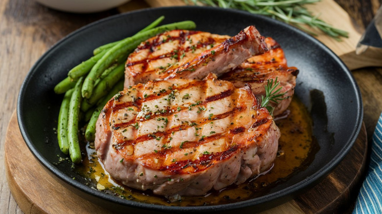 Two pieces of grilled pork chops sit on a bed of sauce in a black plate with a side of green beans.